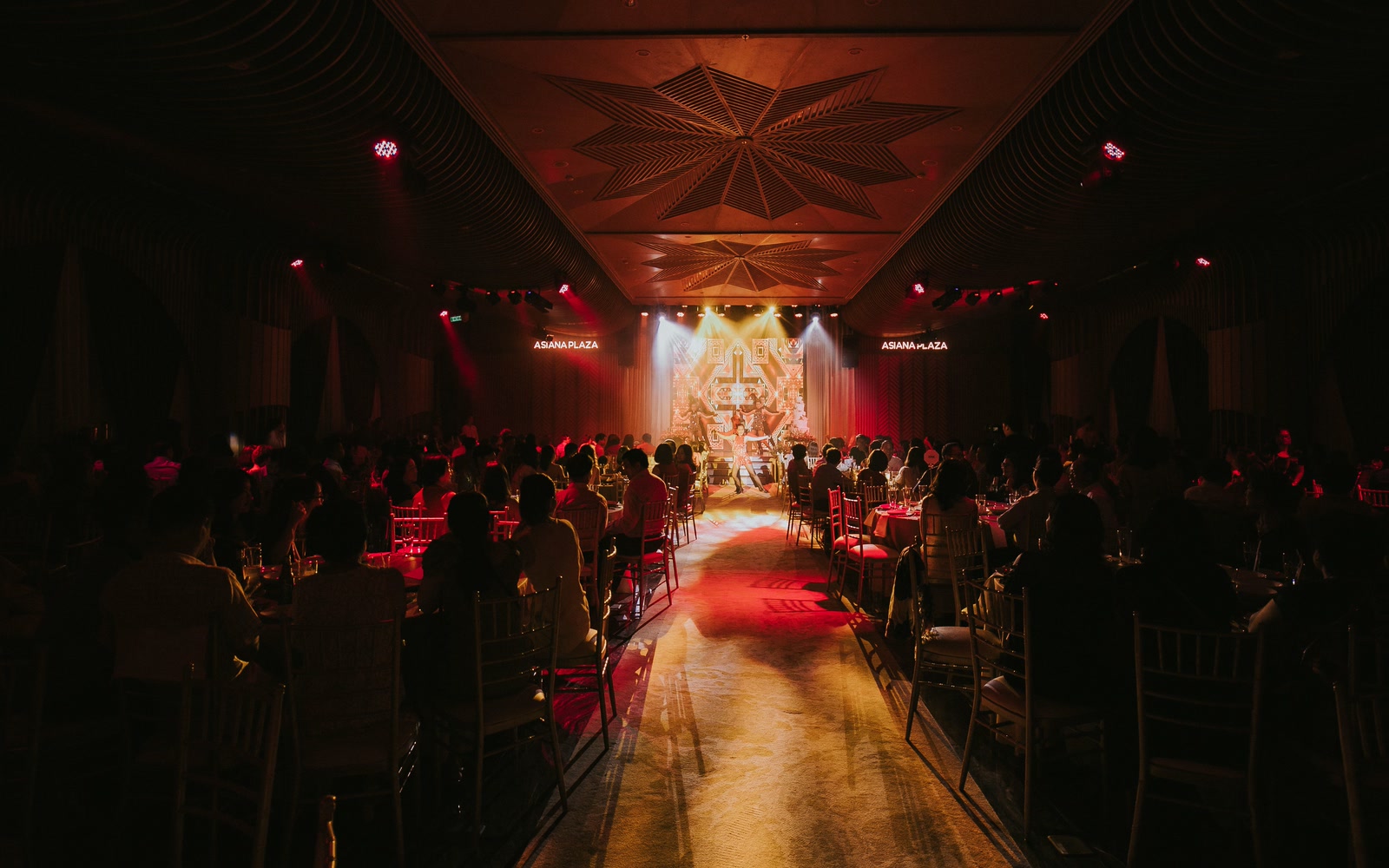 Guests seated at round tables in a banquet hall, facing a lit stage under red uplighting